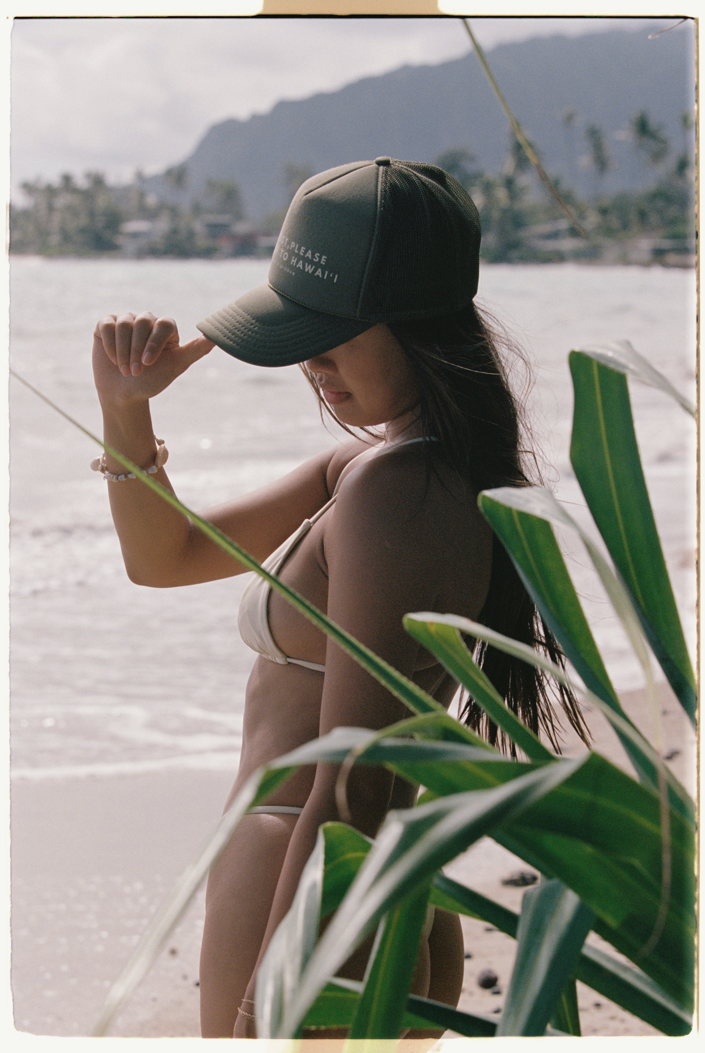 Woman in a bikini and cap sitting on a beach with greenery and water in the background