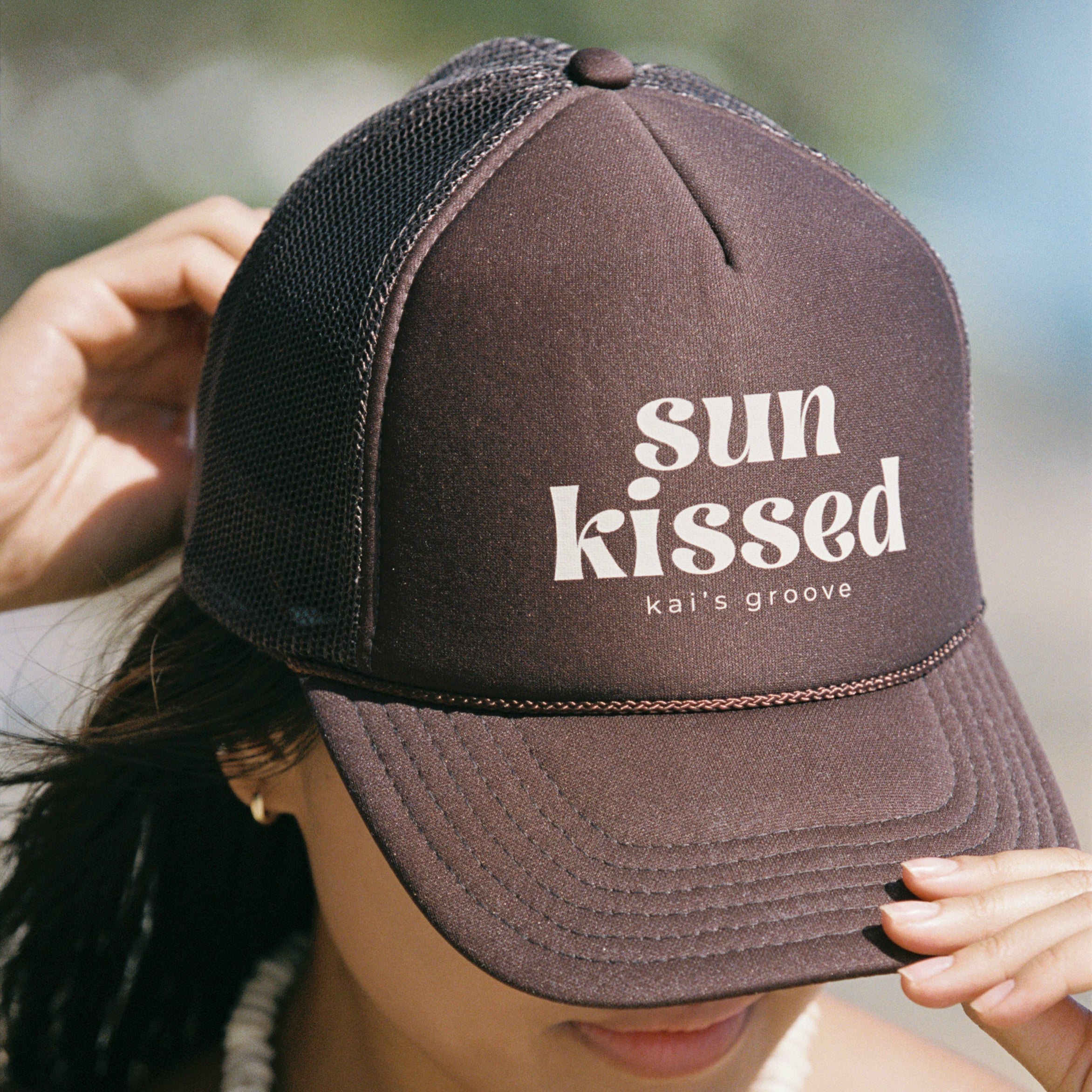 Person wearing a brown cap with 'sun kissed' text, blurred background
