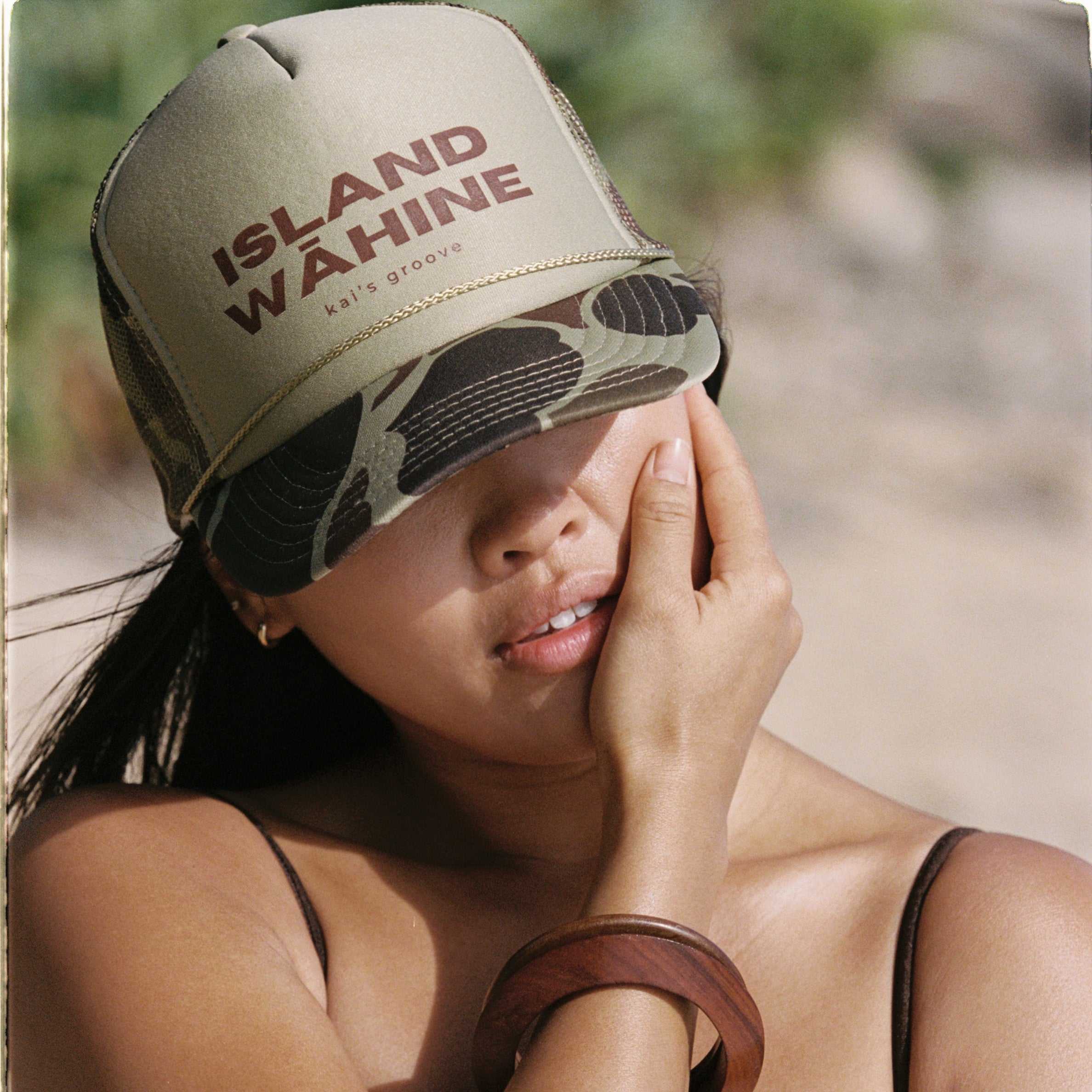 Woman wearing a 'Island Wahine' cap sitting on a beach.