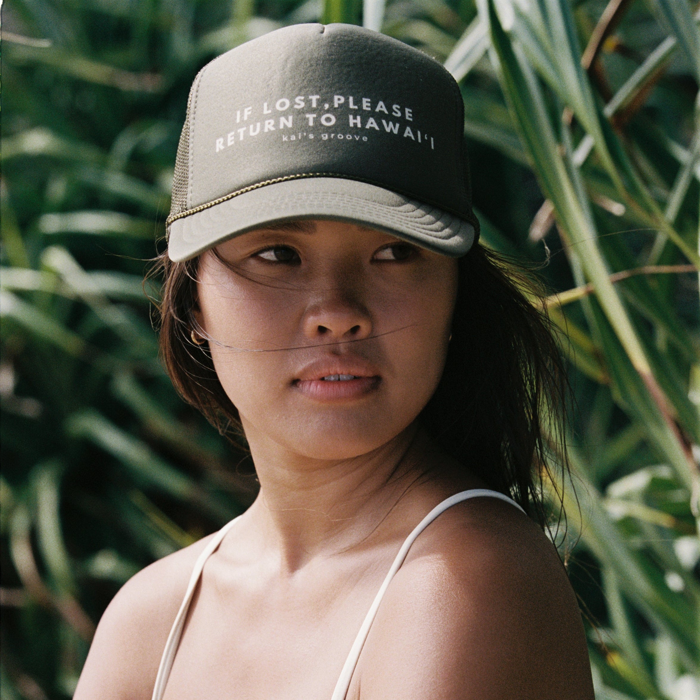 Woman wearing a cap with text and a bikini, standing in front of green foliage.