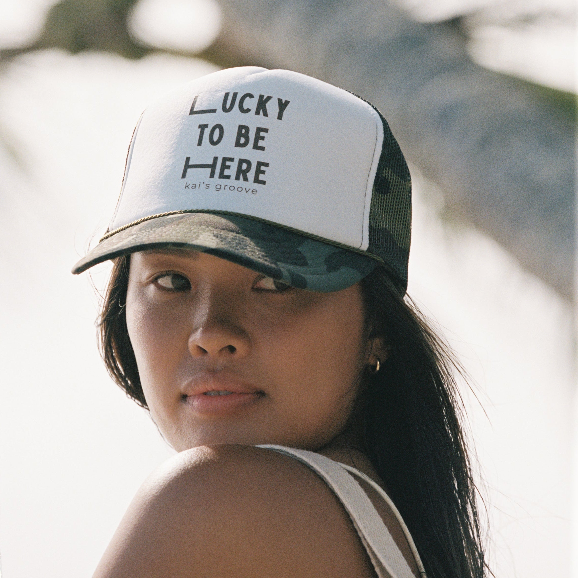 Woman wearing a cap with 'Lucky to be Here' text against a blurred natural background