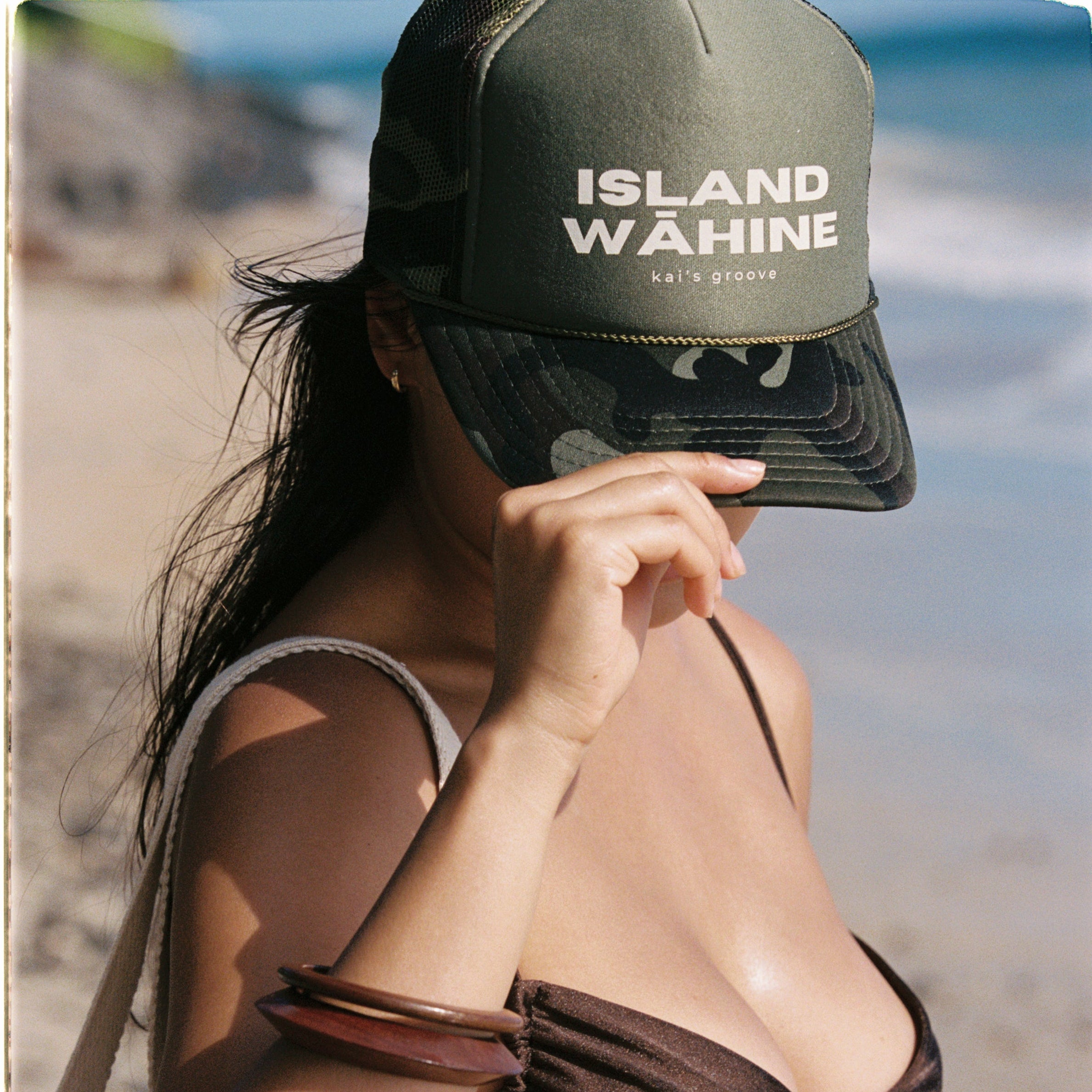 Woman wearing a brown bikini and green camo cap with 'Island Wähine' text on a beach.